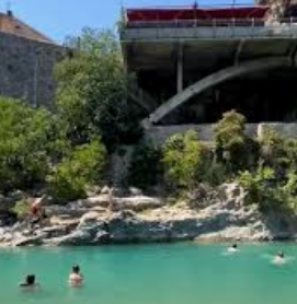 Baigneurs dans une eau turquoise près de rochers et de végétation, avec un pont moderne à l’arrière-plan.