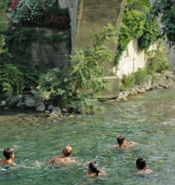 Plusieurs personnes nageant dans la rivière près d’un pilier de pont en pierre entouré de végétation.