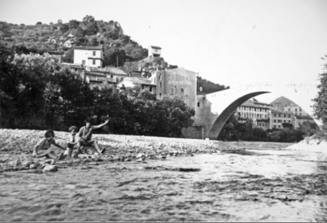 Photographie ancienne de personnes au bord de l’Eygues sur une rive de galets, avec le Pont Roman de Nyons à l’arrière-plan.
