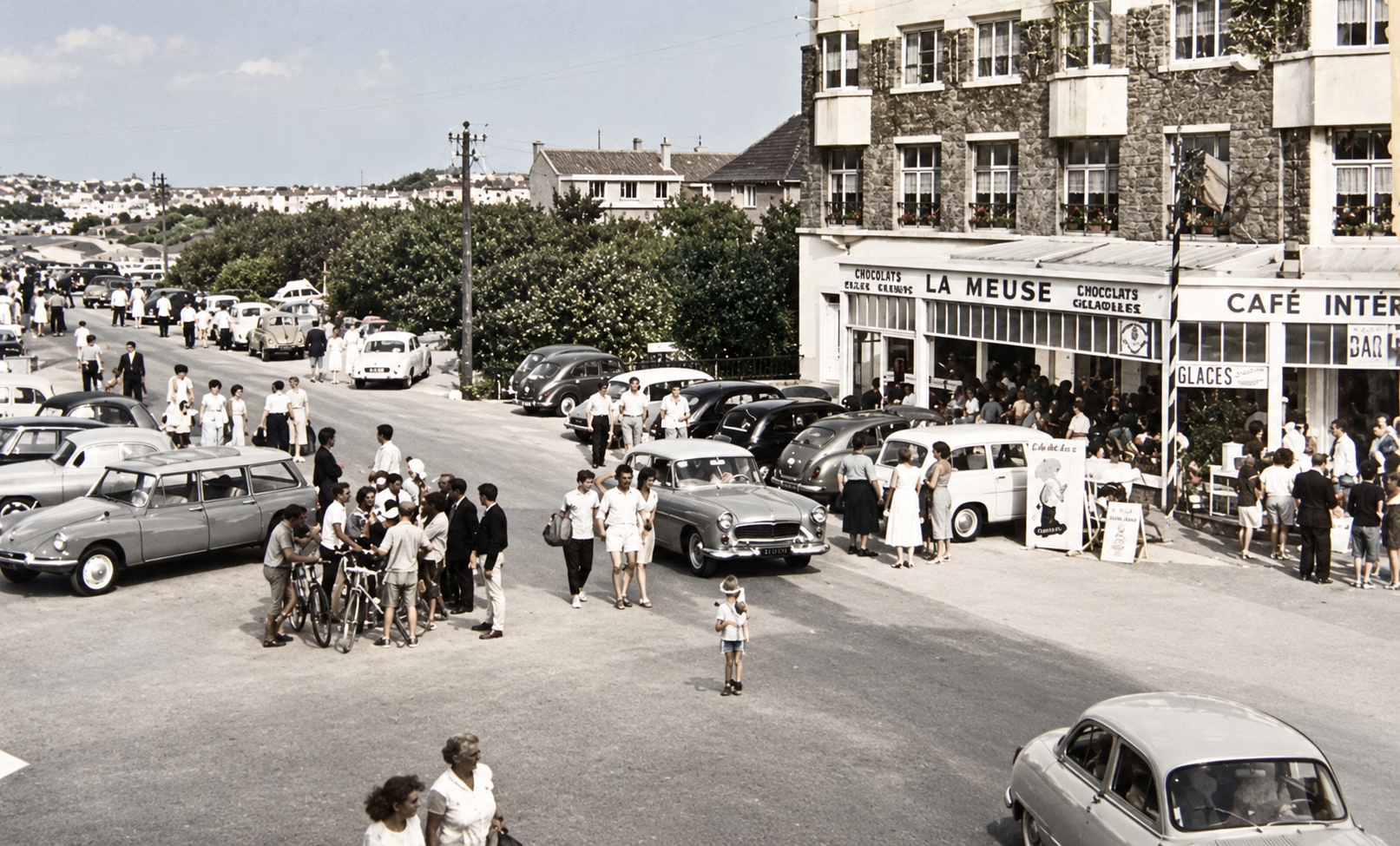 Bray-Dunes vers 1960, rue animée avec voitures anciennes, cyclistes, familles et commerces portant les enseignes La Meuse et Café Inte.