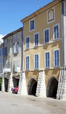 Façade jaune aux volets clairs et arcades médiévales en pierre sur la place du Marché à Buis-les-Baronnies.