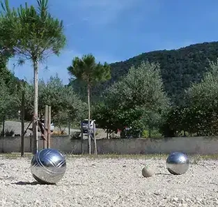Boules et cochonnet sur le terrain de pétanque, avec des arbres et les collines en arrière-plan.