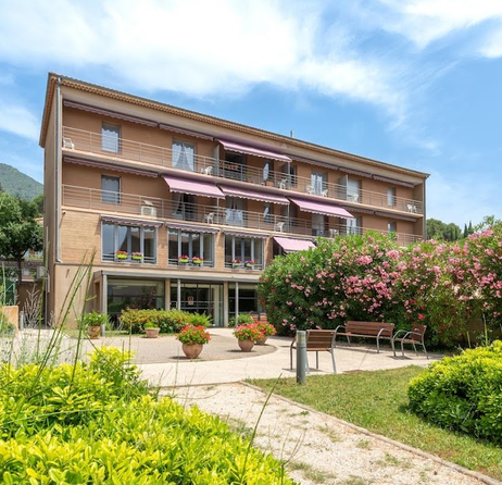 Bâtiment avec balcons et stores roses, entouré d’un jardin avec des bancs.