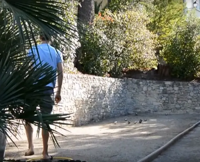 Personne sur un terrain de pétanque bordé d’un mur en pierre et de végétation.