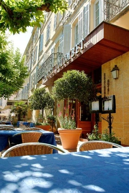 Terrasse de l’Hôtel Colombet à Nyons, avec des tables sous les arbres et une façade aux volets bleus.