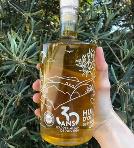 Bouteille d’huile d’olive de Nyons AOP portant la mention 30 ans, tenue devant des branches d’olivier.