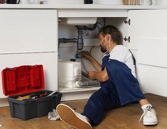 Photo d’illustration d’un plombier travaillant sur les raccordements sous un évier, avec une caisse à outils à côté.