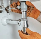 Photo d’illustration de mains gantées manipulant un siphon métallique et les raccords sous un lavabo.