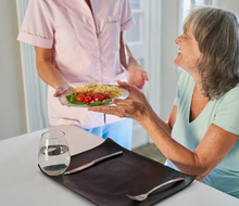 Une personne en tenue rose apporte un plat à une femme âgée assise à une table.
