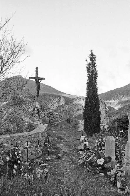 Vue en noir et blanc du cimetière perché de Saint-May, avec tombes fleuries, croix et cyprès devant les montagnes.