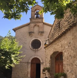 Façade de l’église de Saint-May avec rosace et clocher, derrière les branches d’un arbre et près d’un mur en pierre.