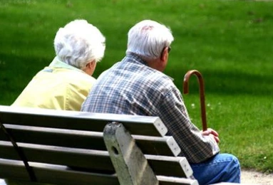 Deux personnes âgées aux cheveux blancs, vues de dos, assises sur un banc face à une pelouse ; l’une tient une canne.
