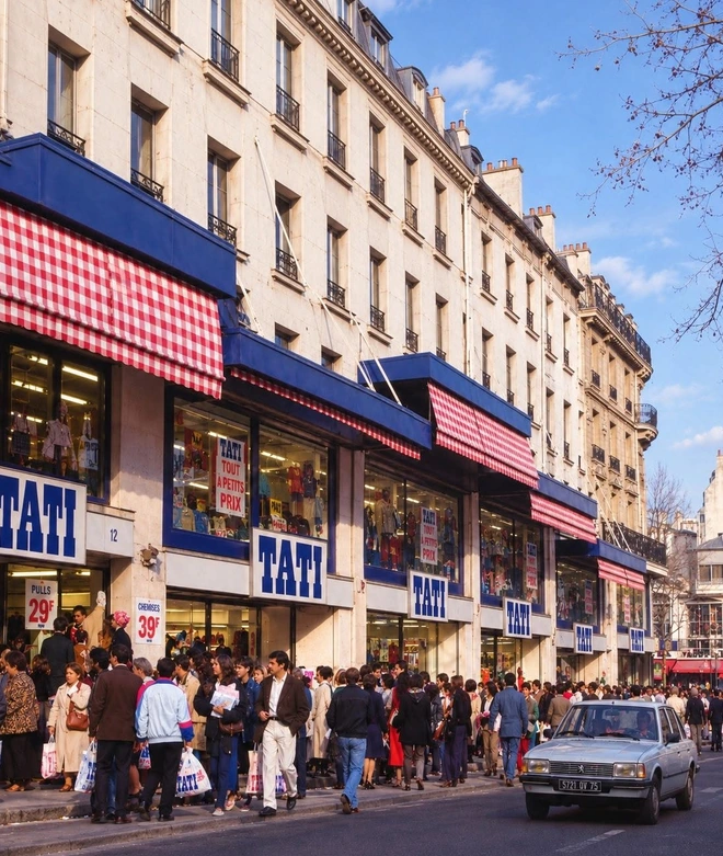 Vue représentant le magasin Tati à Barbès à Paris, ses enseignes bleues, ses auvents à carreaux roses et blancs, ses vitrines et la foule devant les boutiques.