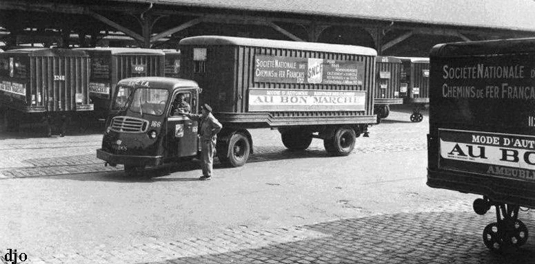 Véhicule routier ancien et remorque portant les inscriptions SNCF et Au Bon Marché dans une cour pavée de marchandises.